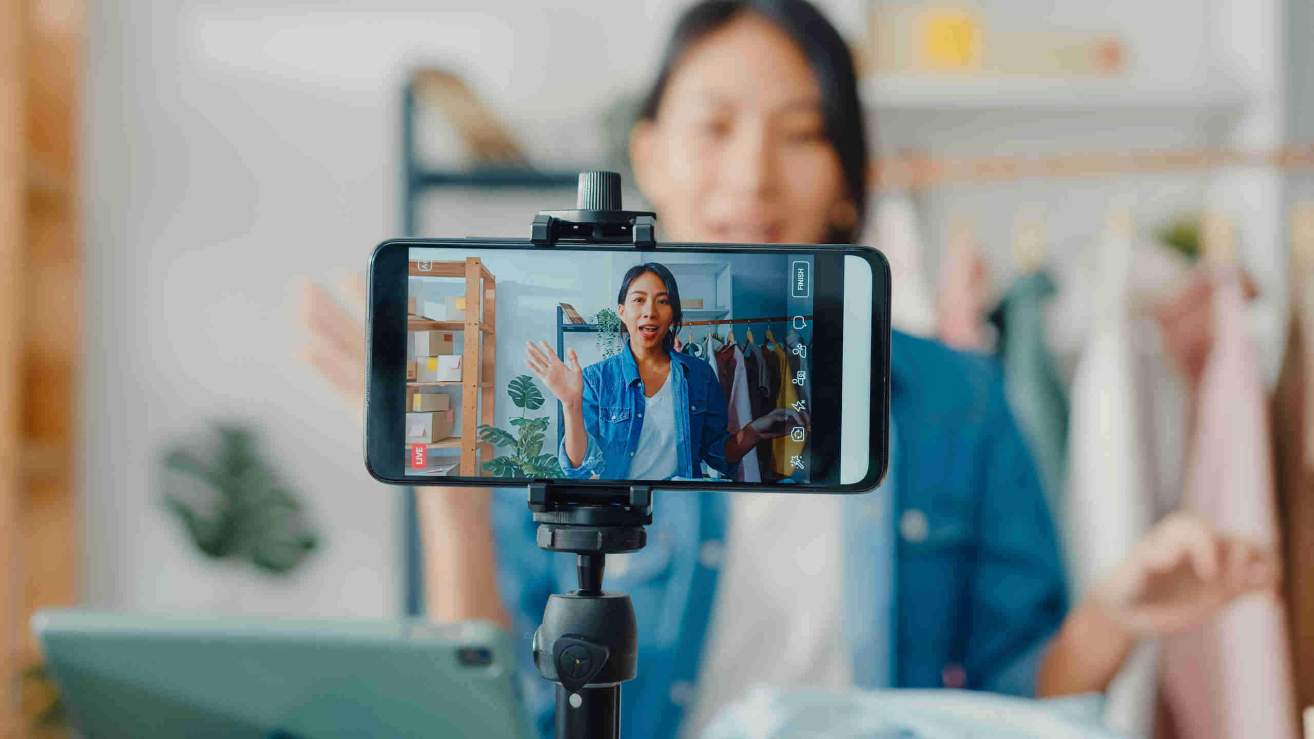 Young Asia lady fashion designer using mobile phone receiving purchase order and showing clothes recording video live streaming online at shop. influencer marketing nyc
